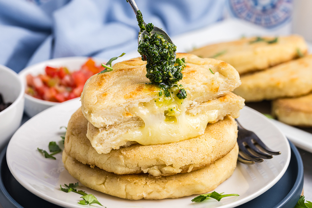 spoon adding chimichurri sauce to stack of arepas